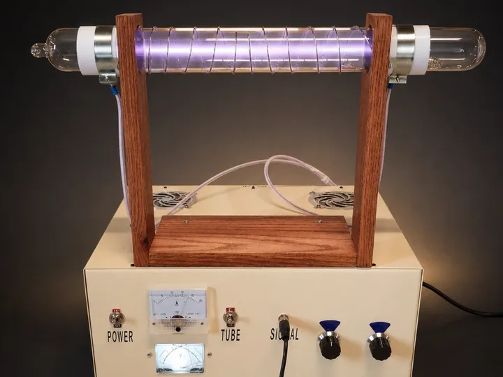 A cream control cabinet with meters below a long violet-glowing plasma tube held on a wooden frame.
